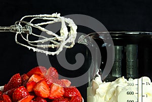 A still life with a bowl of fresh an red strawberries, a food processor, mixer whose wire wisks are full of cream, beside a beaker