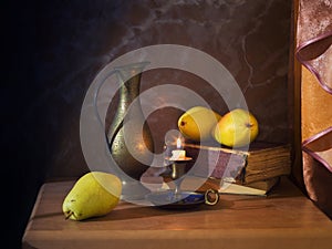 Still life on a brown background, a copper jug, a vintage candlestick, a burning candle, an old book and pears.