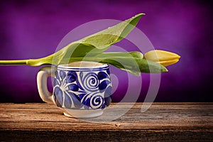 Still life of a closed yellow tulip placed on a blue and white Mexican pottery cup on a barn wood table