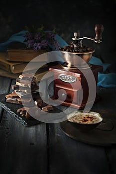 Still life with coffee grinder, stack of chocolate pieces and cup of coffee on wooden table. Dark moody style.