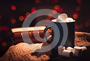 Still life. A cup with marshmallows on a background of colored lights. In the background, a book bound in leather