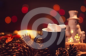 Still life. A cup with marshmallows on a background of colored lights. In the background there is a wreath of spruce and a burning