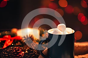 Still life. A cup with marshmallows on a background of colored lights. In the background there is a wreath of spruce and a burning