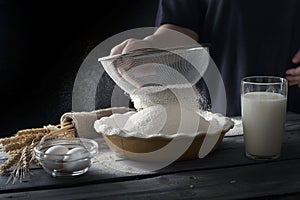 Still life on dark wooden surface. Flour sifting process. Dough ingredients next to bowl with flour.