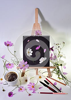 Still life with empty mock up  black sketchbook on a wooden easel, scattered pencils and pens, strewn pink flowers and steaming co