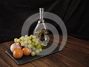 Still life with fresh grapes, tangerines and apple on a slate plate and antique bottle of white wine. Wooden table, dark cloth