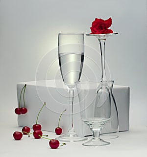 Still life of a glass of water and two empty glasses with a rose and cherries on a white background