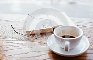 Still life image of cup of black coffee on saucer with vintage book and eyeglasses on the wooden table next to big coffee shop