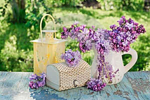 Still-life with a lilac in a jug, a casket, a flashlight on a wooden table