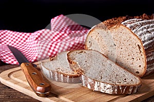 Still life with loaf and sliced wheat and rye bread with knife. On old wooden board with red cloth on black background