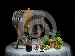 Still life with old oak wine barrel, wine bottle, wine glass and bronze container with bunch of grapes on black background