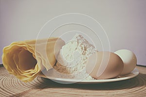 Still life photo, colored eggs, white plate with flour and homemade pasta, on pine wood