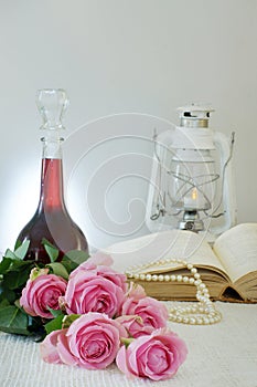 Still life with pink roses, a book, a lantern and a bottle of red wine on a light background