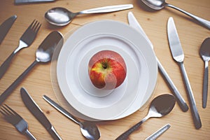 Still Life: An Red Apple Lying on Small Plate. Spoons, Tea Spoons, Forks and Knives on the Wooden Desk as a Background.