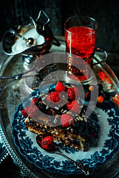 Still life in royal style. Close up piece of chocolate cake with raspberries on old blue plate, beside glass fruit drink on silver