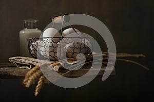 Still life in rustic style. Composition with basket of eggs, bunch of wheat ears on wooden table.