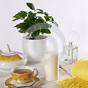 Still life with a standing candle, a cup of tea and a teapot. White wooden table and light background