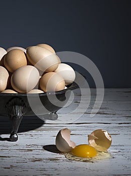 Still life with a tin bowl full of a pile of eggs and a broken egg on a white table with a blue background