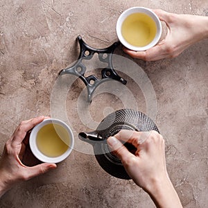 Still life with traditional Asian herbal tea in an old cast-iron teapot with pyalas in hands on a textured background. Square