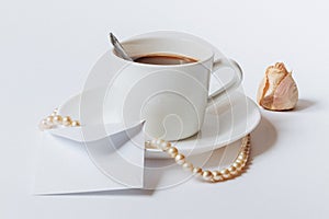 A still life with a white ceramic cup of coffee, a small white envelope, and the dried rose`s bud