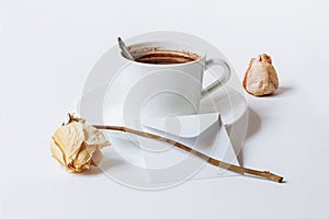 A still life with a white ceramic cup of coffee, a small white envelope, and the dried roses