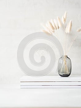 Still life of white dry lagurus flower in glass vase on stack of magazines