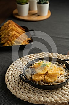 Stir-fried bean sprouts and fried egg tofu sort in black small pan on weaving  placemat, grey placemat, served with snacks, minced