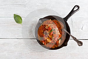 Stir-fried chicken breast in a sauce of tomatoes, garlic, basil and olive oil. Black cast-iron pan, light wooden table, top view