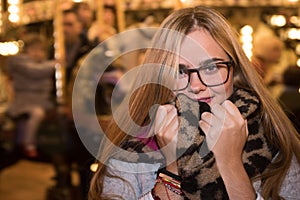 Blonde girl in cold, warms up with scarf on a Christmas night. Carousel in the background