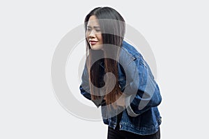 Stomach pain. Side view portrait of beautiful brunette asian young woman in casual blue denim jacket standing and feeling pain on