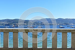 Stone handrail in a beach promenade background. Turqouise water, boats in a bay, blue sky, sunny day. Galicia, Rias Baixas, Spain.