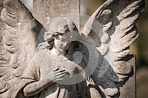 Stone old weathered grave statue of granite weeping angel with broken wing holding chest. Sad peaceful in golden sunshine.