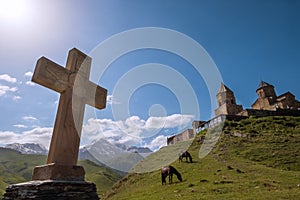 Stone Orthodox cross with a Gergeti Trinity Church with grazing horses on the green grass hill and Kazbek 5054m mountain. Wide