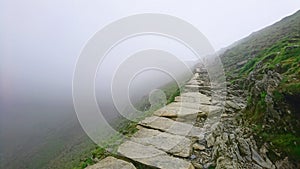 Stone pathway disappearing to vanishing point with drop over edge into fog high up at narrow point on PYG trail on Mount Snowdon i