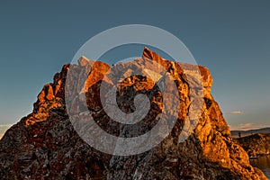 Stone rock in red textured moss in orange bright light of sunset. Cliff illuminated by sun on clear blue sky background. Baikal