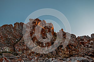 Stone rock in red textured moss in orange bright light of sunset. Cliff illuminated by sun on clear sky background. Baikal