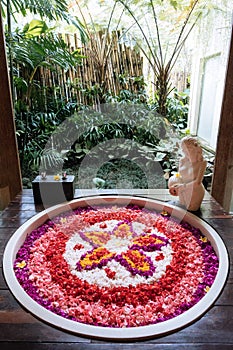 Stone round bath tub with flower shaped petals in pink,red,white colors near window with jungle view. Organic spa