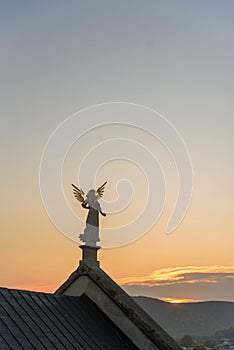 Stone sculpture of an angel with golden wings standing on the rooftop of a baroque temple at sunset.