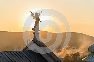 Stone sculpture of an angel with golden wings standing on the rooftop of a baroque temple at sunset.