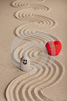 Stone smiley and a red heart stand on a zigzag from the sand.
