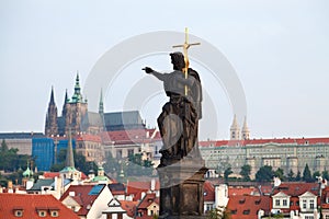 Stone statue on Charles bridge pointing to the St.Vitus Cathedral over red house roofs. Czeck Republick, Prague