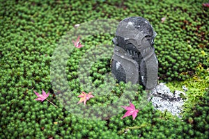 Stone statue of Jizo on ground covered by green star moss and red maple leaves during autumn in a garden at Enkoji temple in Kyoto