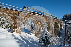 Stone viaduct arch bridge on railway through mountain snowy fir forest and locomotive  with a passenger train. Snow drifts  on