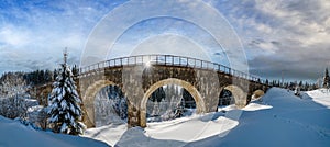 Stone viaduct arch bridge on railway through mountain snowy fir forest. Snow drifts  on wayside and hoarfrost on trees and