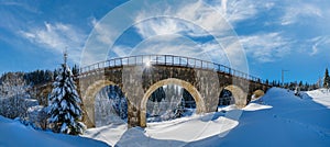 Stone viaduct arch bridge on railway through mountain snowy fir forest. Snow drifts  on wayside and hoarfrost on trees and