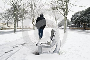 A stone and wood bench covered in snow in the central section of a tree lined street as a man walks by, it is snowing