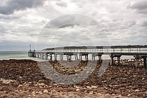 Stones scattered at the shore of Emu Bay with historic jetty during cast cloudy sky on Kangaroo Island in Australia. a