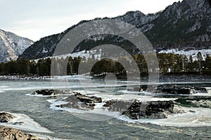 Stony ledges in the middle of the channel of a melted ice-bound river flowing through a mountain valley with snow-capped peaks on