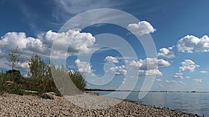 Stony river bank with some shoreline foliage and nice cloudy skies with reflection in water.