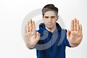 Stop, back off. Serious brunette guy extends palms at camera, telling no, prohibit smth bad, standing over white